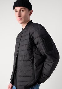 Doudoune noire avec un motif matelassé, poche zippée sur le bras gauche, poignets côtelés et coupe moderne. Portée avec un t-shirt noir en dessous.