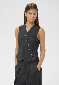 Žena v bezrukávovém tmavě šedém zapínacím vestě a odpovídajících plisovaných kalhotách, s rukama v kapsách, stojící na neutrálním pozadí.