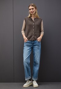 Gilet en tricot marron sur un haut clair, jeans bleu droit avec les revers retroussés, et baskets blanches, sur un fond sombre.