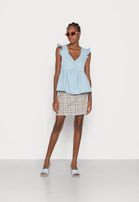 Top peplum azul claro com mangas rendadas, combinado com uma saia mini xadrez. O modelo usa óculos de sol rosa e sandálias azul claro.