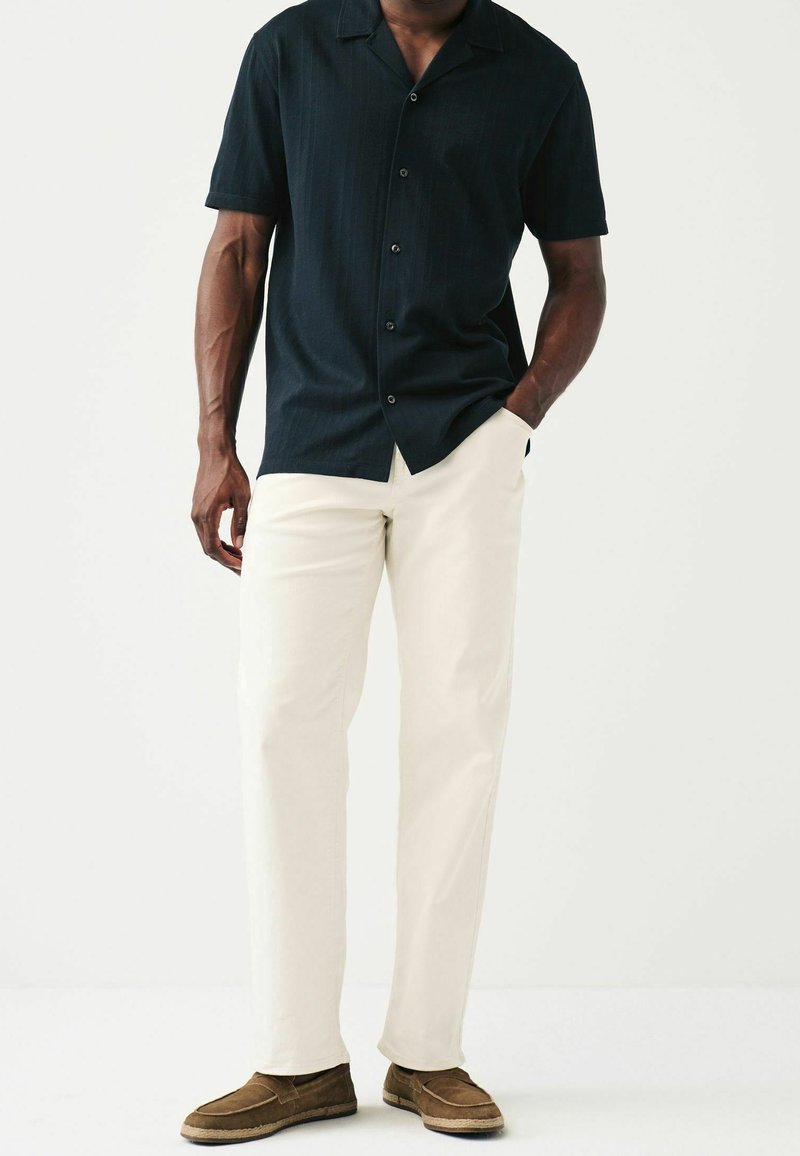 Maglietta nera a maniche corte con una sottile texture, bottoni sul davanti, abbinata a pantaloni color crema e scarpe slip-on marroni.