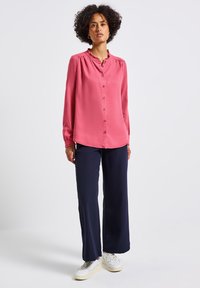 Donna con capelli ricci, che indossa una blusa rosa a maniche lunghe, pantaloni larghi blu scuro e sneakers bianche, in piedi su uno sfondo chiaro e uniforme.