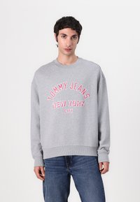 Gray sweatshirt with a crew neck, featuring text "TOMMY JEANS NEW YORK CITY" in pink. Casual fit with ribbed cuffs and waistband.