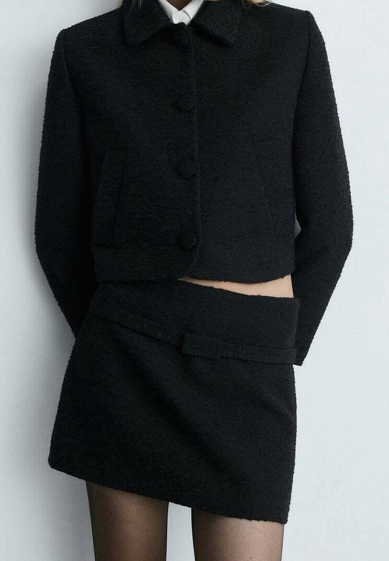 Femme portant une veste courte texturée noire avec une mini-jupe taille haute assortie et des bas noirs transparents sur un fond uni.