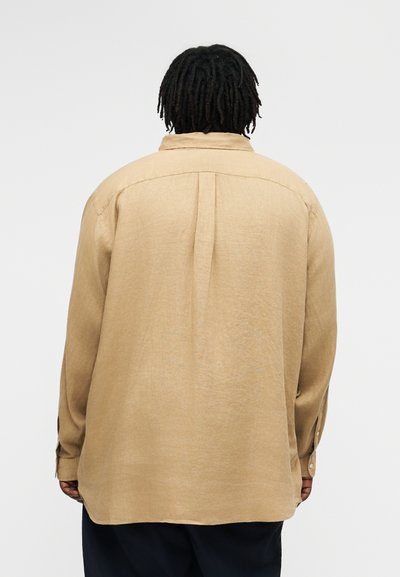 Asmuo su trumpomis dredais, vilkintis laisvą smėlio spalvos ilgomis rankovėmis marškinį ir tamsias kelnes, matomas iš nugaros priešais paprastą foną.