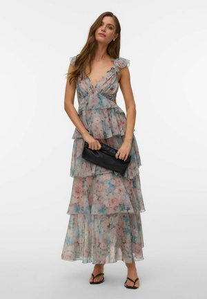 Kobieta ubrana w bezrękawnikową, kwiatową, warstwową sukienkę maxi, trzymająca czarną kopertówkę, stoi z neutralnym wyrazem twarzy na białym tle.