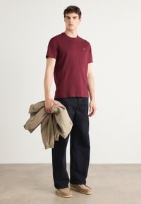 Bordeaux crew-neck t-shirt lavet af bomuld, parret med brede navy bukser og let beige jakke, som holdes i hånden. Suede sko.