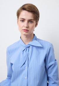 Persona joven con cabello corto castaño claro, lleva una camisa azul con rayas finas y cuello anudado, mirando directamente hacia adelante sobre un fondo blanco liso.