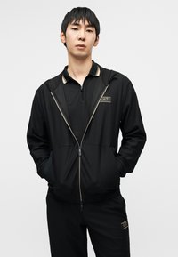 Μαύρο μπουφάν με φερμουάρ, υφασμένο με υφή, μπεζ λεπτομέρειες και πλαϊνές τσέπες, συνδυασμένο με μαύρο πόλο πουκάμισο με μικρό λογότυπο.