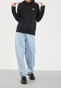 Hoodie noir avec logo jaune, jean bleu clair ample et chaussures noires à lacets. La tenue comprend une poche avant et une capuche ajustable.