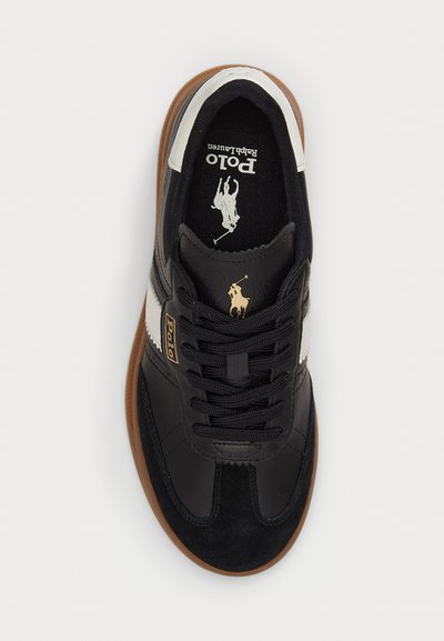 Zwarte sneaker van leer en suede, met een witte streep, zwarte veters en een bruine rubberen zool. Gouden "Polo" logo detail.