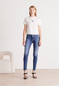 Bílý zkrácený tričko s logem, modré skinny džíny se zesvětlením a černé sandály na popruhy. Neutrální pozadí s světlejším křeslem.