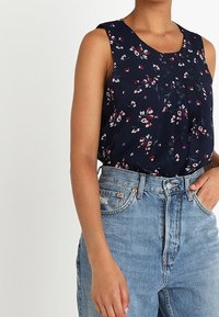 Mörkblå ärmlös blus med blommönster i rött och vitt, med en plisserad front och lättviktig tyg, kombinerad med högmidjade jeans.