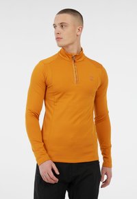 Jaunas vyras, vilkintis prigludusiu oranžiniu užtrauktuku su ilgomis rankovėmis ir juodomis kelnėmis, stovi ir žiūri į šoną, o fone matoma balta spalva.