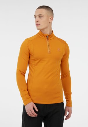 Jonge man in een nauwsluitende oranje lange mouwen zip-up top en zwarte broek, staand en naar de zijkant kijkend tegen een witte achtergrond.