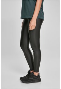 Leggings noirs brillants en matériau extensible, design ajusté, associés à des chaussures de sport noires. Présentés sur un fond neutre.