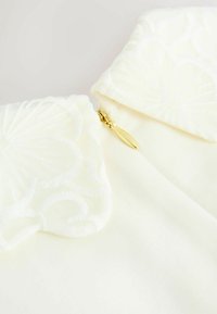 Crème blouse met geborduurde kraag, ritssluiting in goud. Zachte textuur, gelaagde bloemblaadjes in de borduring, elegant en verfijnd ontwerp.