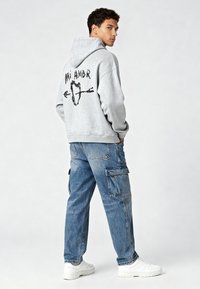 Pilkas džemperis su "MI AMOR" grafika ant nugaros, derinamas su mėlynomis kargo džinsomis ir baltais sportiniais batais. Atsitiktinis, laisvas pjūvis ir minkšta tekstūra.