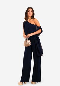 Combinaison en marine à une épaule, design drapé, jambes larges et superposition transparente. Accessoirisée avec un collier doré et un sac beige.