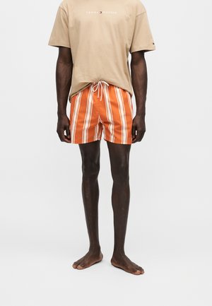 Άνδρας με μπεζ μπλουζάκι Tommy Hilfiger και πορτοκαλί ριγέ μαγιό, όρθιος ξυπόλυτος μπροστά σε απλό λευκό φόντο.