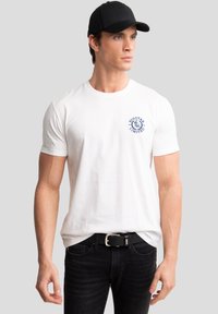 Hvid bomulds-T-shirt med et blåt logo på venstre bryst, parret med sorte jeans og en sort kasket.