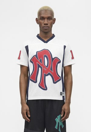 Man with short blonde hair wearing white sports jersey with large red and blue letters and black shorts with green logo, standing facing forward.