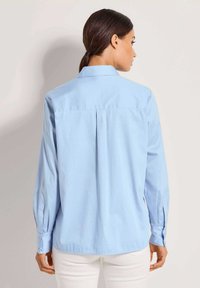 Vrouw met donker haar in een staart, gekleed in een lichtblauw shirt met lange mouwen en een witte broek, met de rug naar de camera gericht tegen een effen achtergrond.