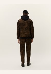 Chaqueta marrón con estampado y pantalones a juego, hechos de tela texturizada, con una capucha y gorra negras, vistos desde la parte trasera.