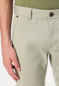 Hellgrüne Baumwollhose mit glatter Textur, ausgestattet mit einem Knopfverschluss und Seitentaschen. Einfaches Design mit dezenten Stichdetails.