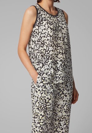 Mujer con camiseta sin mangas negra y pantalones con estampado floral de margaritas blancas, una mano en el bolsillo, sobre fondo gris liso.