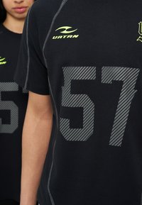 Δύο άτομα φορούν μαύρα αθλητικά μπλουζάκια με γκρι ριγέ αριθμό 57 και κίτρινα λογότυπα, εμφανιζόμενα από το μπροστινό και μερικώς πλάγιο προφίλ.