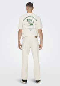 Cream short-sleeve T-shirt with green graphic design on back, paired with beige pants. Wearing black shoes with white sole accents.