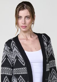 Cardigan a motivi in bianco e nero con una texture lavorata a maglia, caratterizzato da un ampio colletto e design geometrici. Indossato sopra una maglietta bianca.