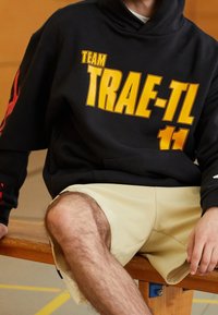 Sweat à capuche noir avec un texte jaune vif "TEAM TRAE-TL 11" sur la poitrine et la manche. Porté avec un short de sport beige, assis sur un banc en bois.