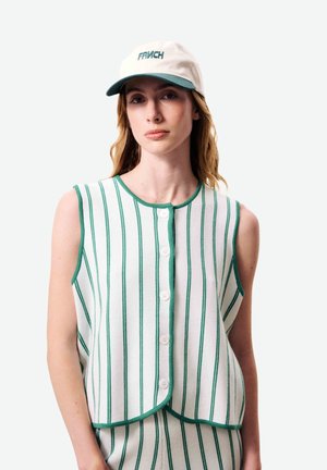 Vrouw draagt een witte mouwloze vest met knopen en bijpassende broek met groene verticale strepen, en een witte pet met groene klep en tekst.