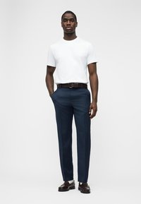 Mann iført hvit T-skjorte, marineblå dressbukser, brunt belte og mørkebrune dressko, stående med hendene i lommene på hvit bakgrunn.