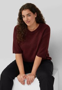 Burgunderfarbener Strickpullover mit kurzen Ärmeln, runden Ausschnitt und weicher Textur. Modell trägt schwarze Hose und sitzt auf einem weißen Würfel.