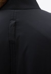 Vue arrière d'une veste noire avec un détail de boucle cousue verticale sous un col côtelé, portée par une personne aux cheveux bruns.
