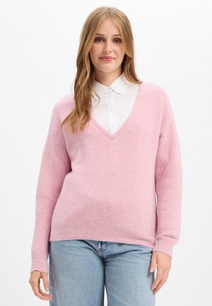 Giovane donna con lunghi capelli biondi che indossa un maglione rosa a scollo a V sopra una camicia bianca con colletto e jeans azzurri chiari su uno sfondo bianco.