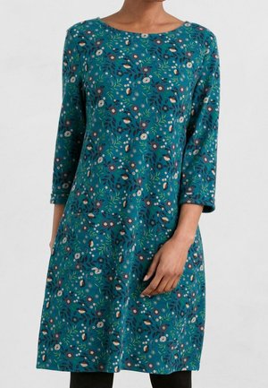 Robe bleu sarcelle avec motif floral et feuilles, manches trois quarts et encolure ronde, portée par une personne avec la main repliée sur le côté.