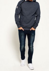 Granatowa bluza z kapturem z kieszenią kangurzą, ściągaczami przy mankietach i zamkiem błyskawicznym na ramieniu, zestawiona z ciemnymi jeansami i białymi trampkami.