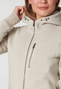 Beige zip-up hoodie med hög krage, justerbar dragsko och framficka med dragkedja. Har texturerad tyg och broderad logotypdetalj.