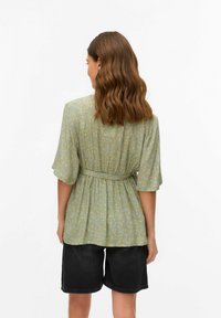 Zelena cvetlična bluzica s kratkimi, širokimi rokavi, nabrano pasom in mehkim teksturo, v kombinaciji s temnimi hlačami iz denima. Prikazan zadnji pogled.