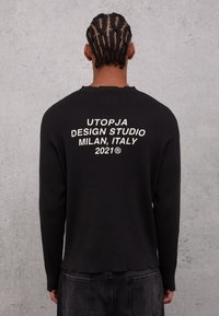 Maglione nero a costine con collo a girocollo, caratterizzato da ricami bianchi sulla schiena che fanno riferimento a "Utopja Design Studio, Milano, Italia 2021."