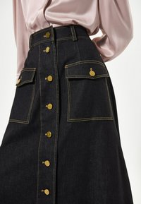 Gonna di jeans blu scuro con bottoni dorati, vita alta, due tasche frontali con alette e dettagli di cuciture gialle a contrasto.