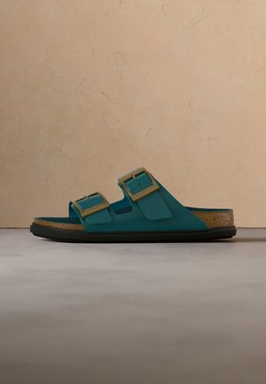 Turkusowa sandałka typu slide z dwoma złotymi klamrami i korkową wkładką na jednolitym beżowym tle.