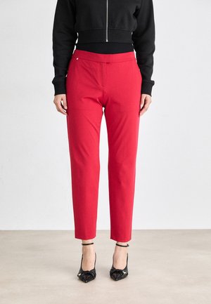 Pantalons rouges ajustés avec une coupe droite, fermeture éclair et tissu lisse ; associés à des chaussures à talons hauts noires et une veste noire courte.