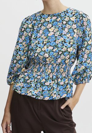Frau trägt eine Bluse mit Blumenmuster in Blau, Rosa und Gelb, mit geraffter Taille und gepufften Dreiviertelärmeln.