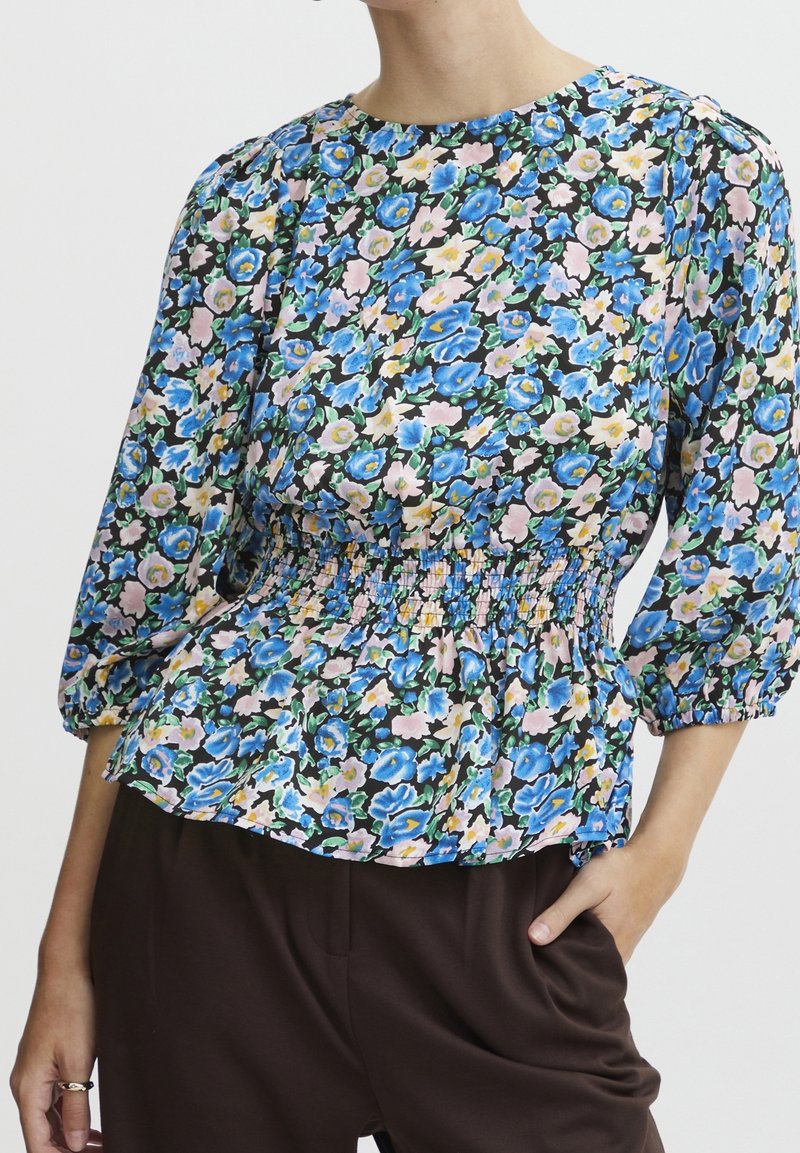Femme portant un chemisier à fleurs bleues, roses et jaunes, avec une taille froncée et des manches trois-quarts bouffantes.