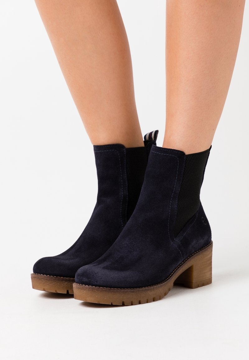 Bottes en daim bleu marine montant jusqu'à la cheville avec talons carrés et semelles en caoutchouc marron, portées sur jambes nues sur un fond blanc.